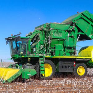 May include: A large green and yellow cotton picker machine in a cotton field under a clear blue sky. The machine has large yellow wheels and a cylindrical bale of cotton. The text "C5690" is visible on the side.
