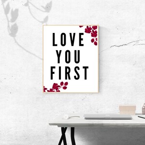 Puede incluir: Una impresión enmarcada con fondo blanco y texto negro que dice "LOVE YOU FIRST". Ilustraciones florales rojas se encuentran en los lados izquierdo y derecho del texto.