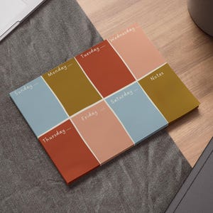 Pode incluir: Bloco de notas de planejamento semanal com um design geométrico. O bloco de notas apresenta blocos de cores azul, amarelo mostarda, coral e pêssego, com os dias da semana escritos em branco. O planejador inclui uma seção de notas.