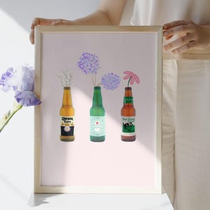 Puede incluir: Impresi&oacute;n enmarcada que presenta tres botellas de cerveza que se utilizan como jarrones para flores. Las botellas son Corona Extra, Heineken y New Glarus. Las flores son hortensias moradas y una margarita rosa. El fondo es rosa claro.