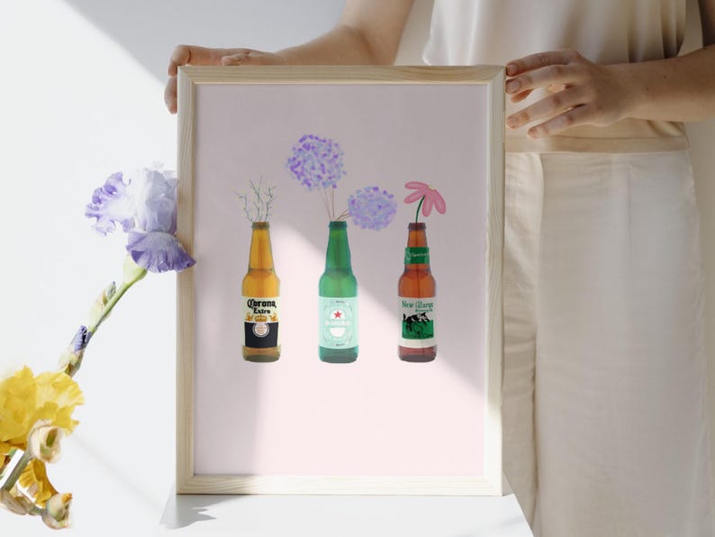 Puede incluir: Impresi&oacute;n enmarcada que presenta tres botellas de cerveza que se utilizan como jarrones para flores. Las botellas son Corona Extra, Heineken y New Glarus. Las flores son hortensias moradas y una margarita rosa. El fondo es rosa claro.
