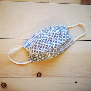 May include: A handmade face mask with a white and navy blue geometric pattern. The mask has white elastic ear loops and is laying on a light wood surface. The mask is designed to cover the nose and mouth.