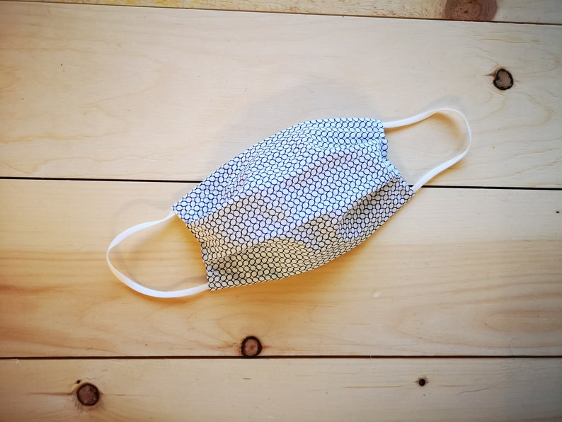 May include: A handmade face mask with a white and navy blue geometric pattern. The mask has white elastic ear loops and is laying on a light wood surface. The mask is designed to cover the nose and mouth.