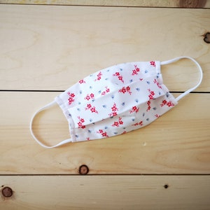 May include: White face mask with a floral pattern of red flowers and small blue accents. The mask has white elastic ear loops and is made of a soft, pleated fabric. The mask is designed for protection and comfort.