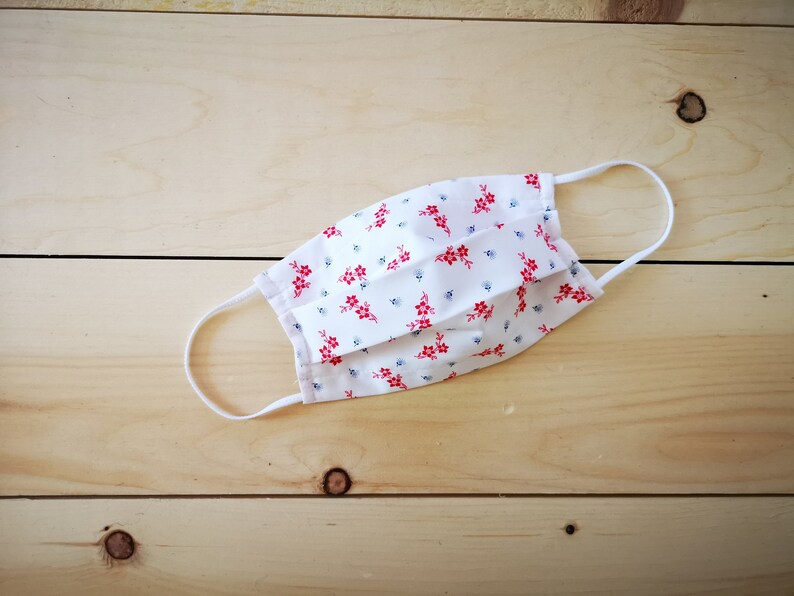 May include: White face mask with a floral pattern of red flowers and small blue accents. The mask has white elastic ear loops and is made of a soft, pleated fabric. The mask is designed for protection and comfort.