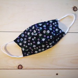 May include: A black face mask with a floral pattern of white, purple, and green flowers. The mask has white elastic ear loops and is made of fabric. The mask is designed to cover the nose and mouth.