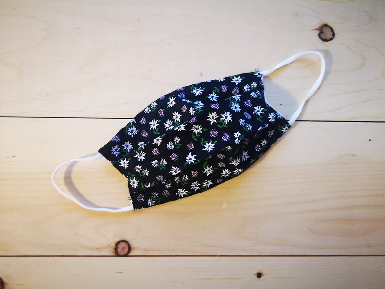 May include: A black face mask with a floral pattern of white, purple, and green flowers. The mask has white elastic ear loops and is made of fabric. The mask is designed to cover the nose and mouth.