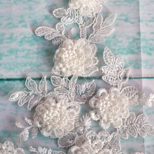 May include: Close-up of white floral lace with raised, textured flowers and delicate leaf patterns. The lace is set against a light blue and white background, creating a soft, elegant aesthetic. Ideal for bridal or craft projects.