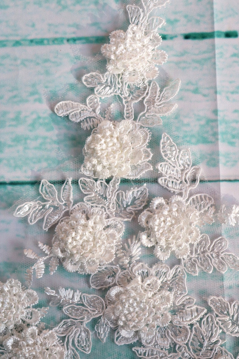 May include: Close-up of white floral lace with raised, textured flowers and delicate leaf patterns. The lace is set against a light blue and white background, creating a soft, elegant aesthetic. Ideal for bridal or craft projects.