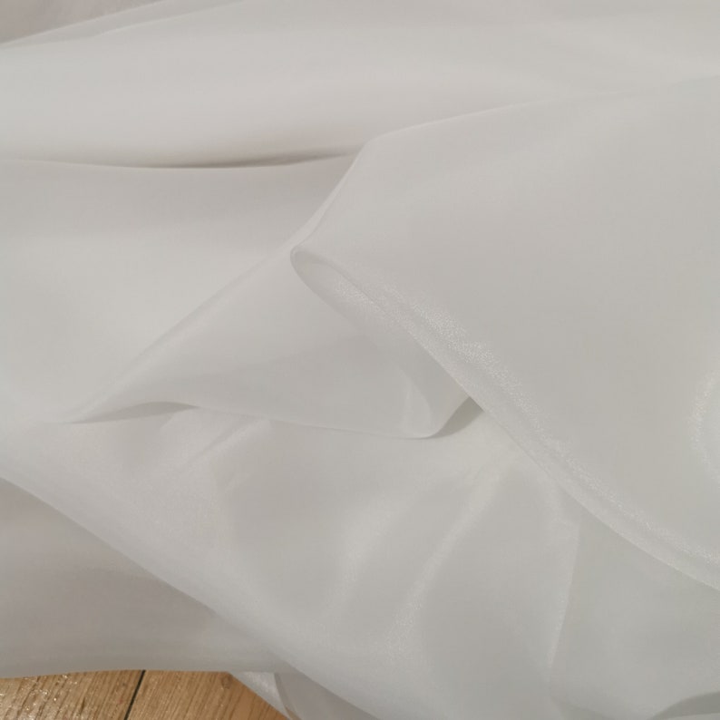 May include: White sheer fabric with a soft, flowing texture. The fabric is folded and draped, showcasing its delicate and translucent quality.