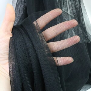 Puede incluir: Primer plano de tela de tul negra. El material transparente se sostiene, revelando su delicada estructura en forma de red. La tela es de color negro intenso, con una apariencia suave y fluida. Ideal para manualidades, moda y proyectos decorativos.