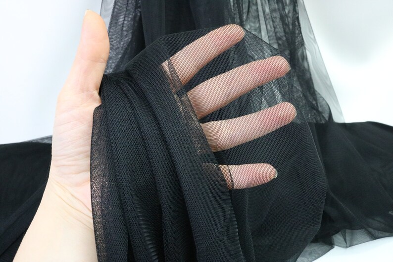Puede incluir: Primer plano de tela de tul negra. El material transparente se sostiene, revelando su delicada estructura en forma de red. La tela es de color negro intenso, con una apariencia suave y fluida. Ideal para manualidades, moda y proyectos decorativos.