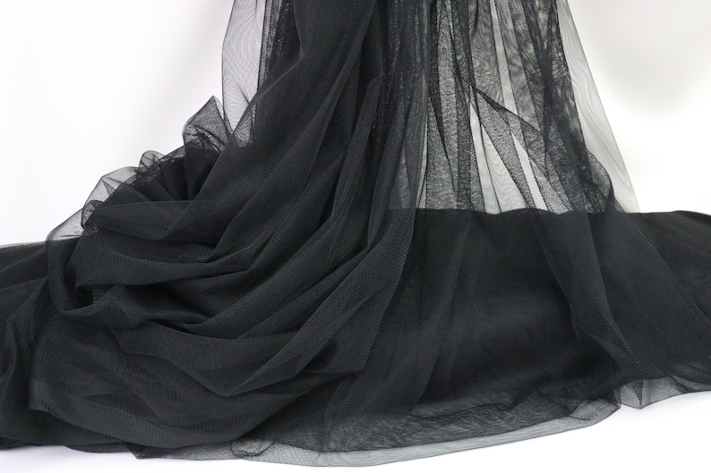 Puede incluir: Tela de tul negra, drapeada y en capas, creando un efecto fluido y texturizado. El material transparente est&aacute; recogido y doblado, con luz que pasa a trav&eacute;s del delicado tejido. La tela est&aacute; sobre un fondo blanco.
