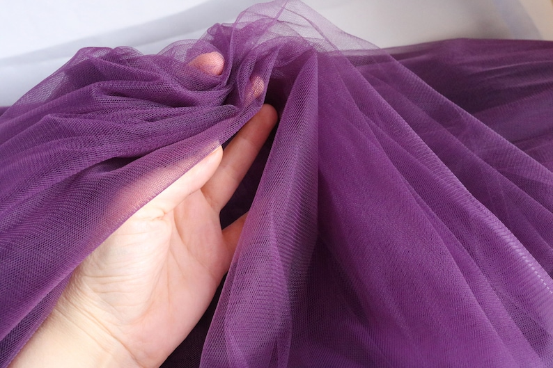 May include: Purple tulle fabric with a soft, delicate texture. The fabric is sheer and has a fine mesh weave.