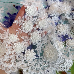 May include: Ivory lace fabric with floral and leaf embroidery. The delicate lace features three-dimensional, clustered flower embellishments. The lace has a scalloped edge and is suitable for bridal wear, crafts, or other decorative projects.