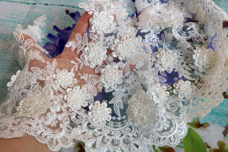 May include: Ivory lace fabric with floral and leaf embroidery. The delicate lace features three-dimensional, clustered flower embellishments. The lace has a scalloped edge and is suitable for bridal wear, crafts, or other decorative projects.