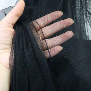 Puede incluir: Primer plano de tela de tul negra. El material transparente se sostiene en una mano, mostrando su delicada estructura de red. La tela es opaca y parece estar en capas, creando profundidad y textura. El fondo es blanco suave.