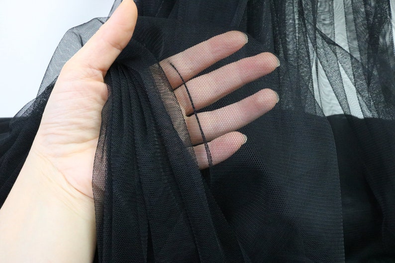 Puede incluir: Primer plano de tela de tul negra. El material transparente se sostiene en una mano, mostrando su delicada estructura de red. La tela es opaca y parece estar en capas, creando profundidad y textura. El fondo es blanco suave.