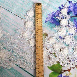 May include: Ivory lace trim with floral and leaf designs, featuring raised, textured flower details. A wooden ruler shows measurements in inches. The lace is set against a light blue and white background with purple flowers.