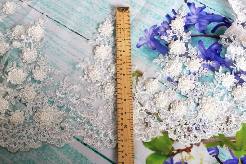 May include: Ivory lace trim with floral and leaf designs, featuring raised, textured flower details. A wooden ruler shows measurements in inches. The lace is set against a light blue and white background with purple flowers.
