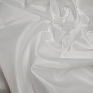 Puede incluir: Tela blanca con una textura suave y sedosa. La tela está drapeada y doblada, creando curvas suaves y fluidas.