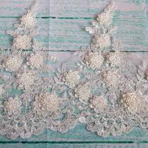 May include: White floral lace trim with 3D flower and pearl accents. The lace features a delicate, openwork design with detailed floral patterns and scalloped edges. Ideal for bridal wear, crafts, or embellishments.