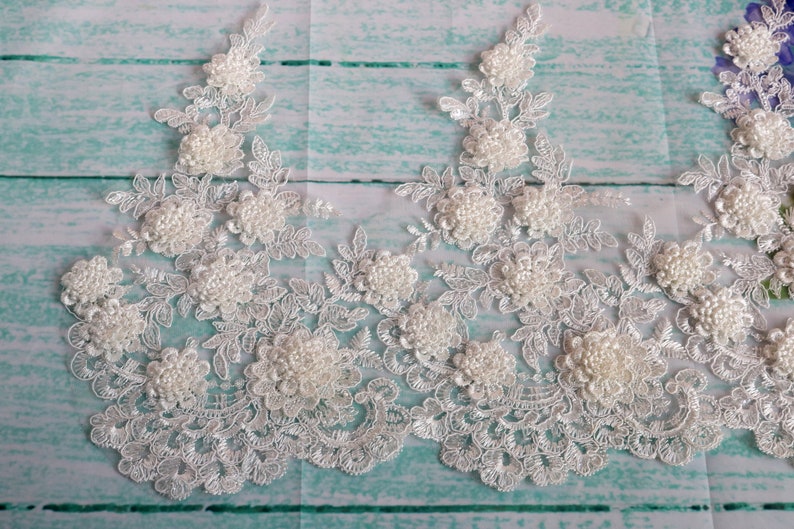 May include: White floral lace trim with 3D flower and pearl accents. The lace features a delicate, openwork design with detailed floral patterns and scalloped edges. Ideal for bridal wear, crafts, or embellishments.