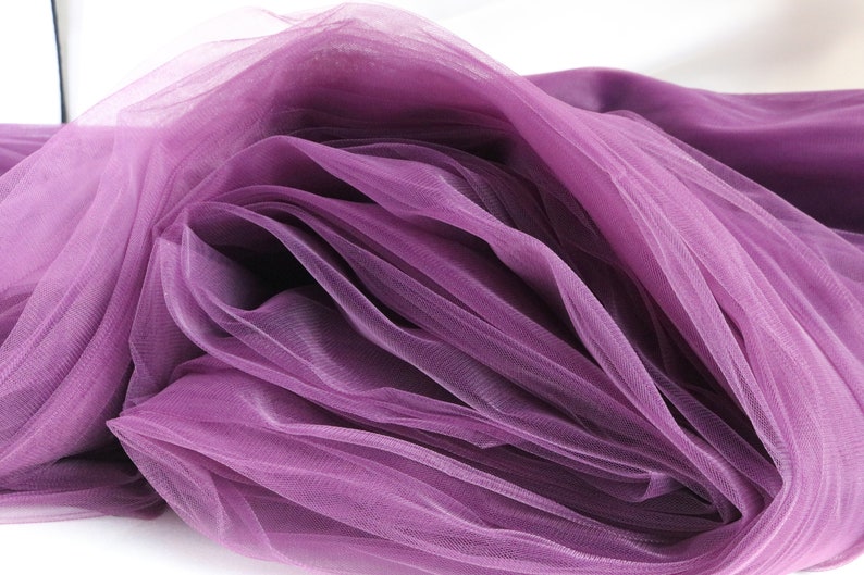 May include: Purple tulle fabric with a soft, delicate texture. The fabric is folded and draped, showcasing its sheer and airy quality.