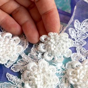 May include: Close-up of white floral lace applique. The applique features detailed, three-dimensional flower designs with delicate, embroidered leaves. The lace is set against a blue and purple background.