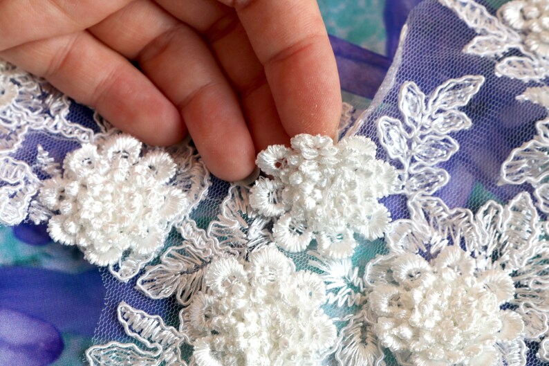 May include: Close-up of white floral lace applique. The applique features detailed, three-dimensional flower designs with delicate, embroidered leaves. The lace is set against a blue and purple background.
