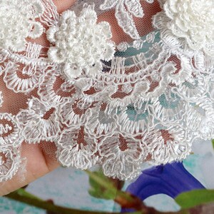 May include: Close-up of white lace fabric with intricate floral and scrollwork embroidery. The lace features raised, textured floral appliques and delicate scalloped edges, perfect for bridal or craft projects.