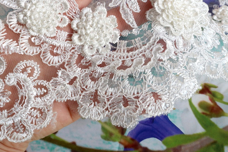 May include: Close-up of white lace fabric with intricate floral and scrollwork embroidery. The lace features raised, textured floral appliques and delicate scalloped edges, perfect for bridal or craft projects.