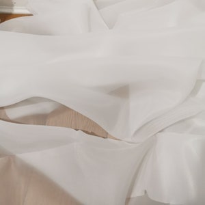 May include: White sheer fabric with a smooth texture. The fabric is folded and draped, creating soft folds and curves.