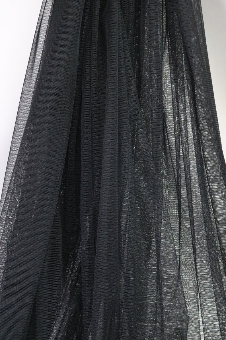 Puede incluir: Primer plano de tela de tul negra. La tela est&aacute; drapeada y tiene un tejido fino y abierto, creando un efecto semitransparente. Los pliegues y las sombras a&ntilde;aden profundidad y textura al material.