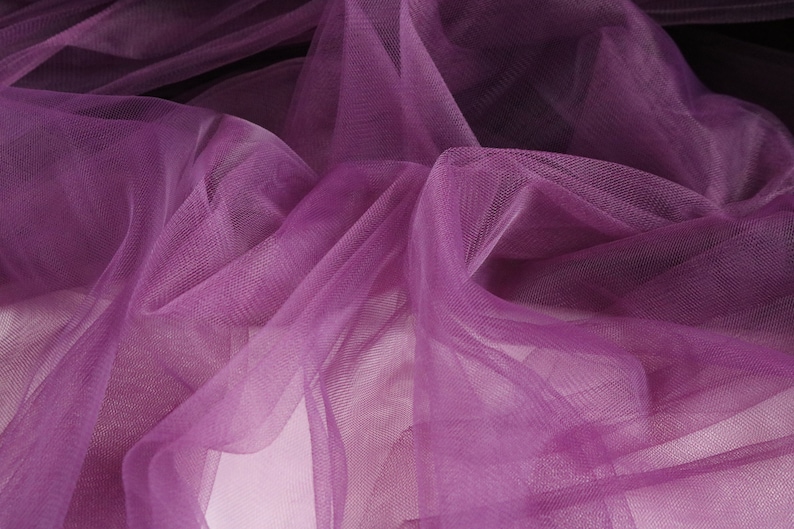 May include: Purple tulle fabric with a soft, delicate texture. The fabric is draped and folded, creating a flowing and ethereal look.