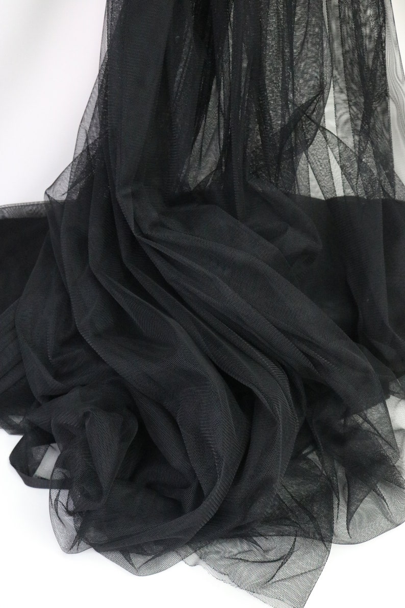 Puede incluir: Primer plano de tela de tul negra. La tela est&aacute; drapeada y doblada, creando sombras y texturas. El material parece ligero y transparente, con un tejido de malla fina. El fondo es blanco.
