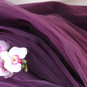 May include: Three white orchid flowers with pink centers are arranged on a purple tulle fabric.
