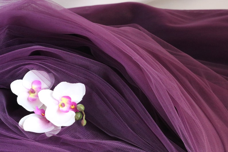 May include: Three white orchid flowers with pink centers are arranged on a purple tulle fabric.