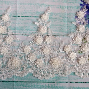 May include: Ivory lace trim featuring floral and beaded embellishments. The delicate lace has a scalloped edge and is adorned with clusters of small, round beads, creating a textured, three-dimensional effect. Ideal for bridal wear or craft projects.