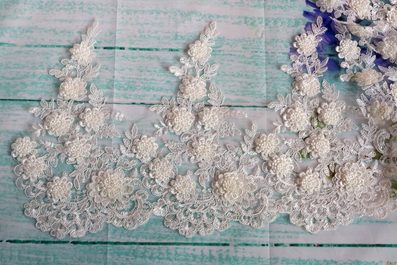 May include: Ivory lace trim featuring floral and beaded embellishments. The delicate lace has a scalloped edge and is adorned with clusters of small, round beads, creating a textured, three-dimensional effect. Ideal for bridal wear or craft projects.