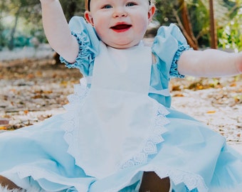 May include: A baby girl dressed as Alice in Wonderland, wearing a blue dress with a white apron and a black bow headband. She is sitting on a white fluffy rug with a happy expression on her face.