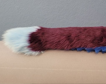 May include: A faux fur tail in shades of burgundy, blue, and light blue. The tail is long and cylindrical, with a fluffy texture. The base is a vibrant blue, transitioning to burgundy, and ending with a light blue tip.