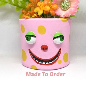 May include: A pink ceramic planter with a whimsical face, featuring yellow polka dots, green eyes, and a wide smile. The planter is filled with artificial flowers in shades of pink, yellow, and green. The text "Made To Order" is printed below the planter.
