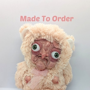 May include: A pink plush teddy bear with a human-like face and large, green eyes. The bear is wearing a pink ribbon around its neck and has a "Made To Order" text label above it.