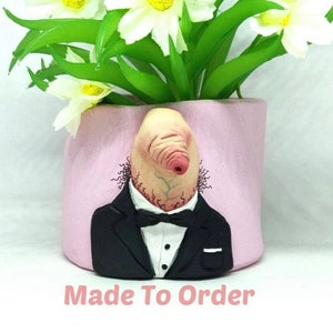 May include: A pink ceramic planter shaped like a person's head with a nose and a bow tie. The planter has a white shirt and a black suit jacket. The planter is holding a bouquet of white flowers.
