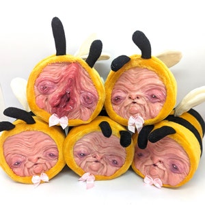 May include: Five plush bee toys with human-like faces. The bees are yellow and black with pink bows. The bees have realistic-looking skin and eyes.