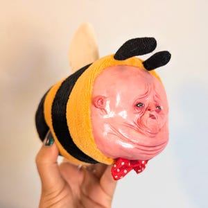 May include: A plush bee toy featuring a soft yellow and black striped body with two black antennae and a single wing. The face is a realistic, textured, flesh-toned sculpture with detailed eyes, nose, and mouth, accented by a small red polka-dot bow tie.