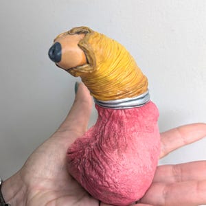 May include: A sculpted figurine featuring a textured, pink, rounded base connected by a silver-coloured ring to a yellow, ribbed, cylindrical upper section that terminates in a smooth, tan, rounded tip with a small, dark, circular indentation.