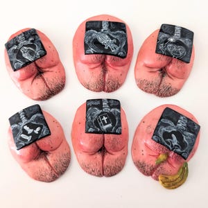May include: A set of six sculpted pink buttocks-shaped magnets, each featuring a black square insert displaying a white line-art illustration, including a troll doll, an alien, a book with a cross, and a banana, with visible hair details.