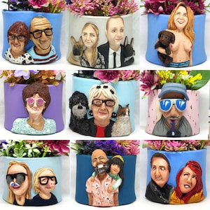 May include: A collection of handmade ceramic planters, each featuring a unique sculpted portrait of a person or couple. The planters are painted in various colors and filled with vibrant pink and purple flowers. Each planter is approximately 5 inches tall.